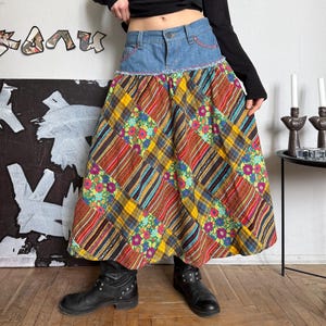 May include: A long, patchwork skirt with a denim waistband and a colorful plaid and floral pattern. The skirt is paired with a black long-sleeve top and black boots. The skirt features a mix of textures and colors.