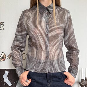 May include: A long-sleeved button-down shirt featuring a semi-sheer fabric with a swirling gray and brown marble-like pattern. The shirt has a classic collar, a front button placket with small dark buttons, and straight cuffs on the sleeves.