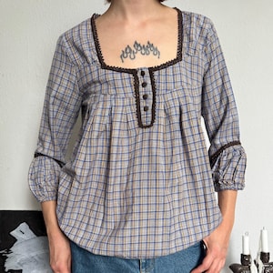 Pode incluir: Uma blusa de manga comprida com padrão xadrez azul, amarelo e branco, com decote quadrado, acabamento em croché castanho e quatro pequenos botões castanhos. A blusa tem mangas franzidas com acabamento castanho e um corte largo e fluido.