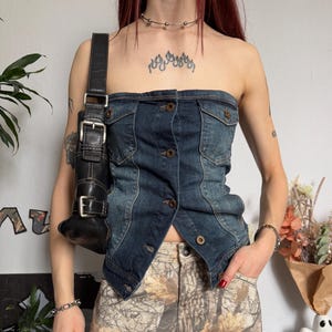 Puede incluir: Un top de mezclilla sin tirantes con botones y bolsillos se combina con pantalones con estampado de camuflaje. Un bolso de hombro de cuero negro con hebillas plateadas se lleva sobre el hombro. El atuendo se complementa con una gargantilla y una pulsera plateadas.