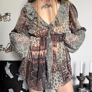 Può includere: Abito fantasia con maniche lunghe e scollo a V. L'abito presenta un mix di motivi paisley e floreali nelle tonalità del marrone, blu e crema. Ha un colletto a balze e un fiocco in vita, creando uno stile bohémien.
