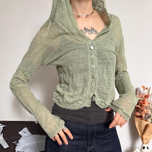 Può includere: Un cardigan con cappuccio color verde salvia, trasparente e a maniche lunghe, caratterizzato da un'abbottonatura frontale con piccoli bottoni circolari iridescenti e un orlo grezzo ondulato. Il capo è indossato sopra una canotta grigio scuro e jeans blu scuro.