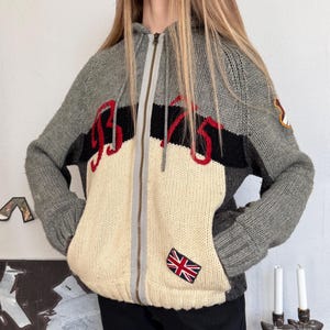 May include: A gray, cream, and black zip-up hooded sweater with the word "9375" in red lettering across the front. Features a Union Jack patch and a small patch on the sleeve. The sweater has a relaxed fit and two front pockets.