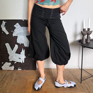 May include: A short-sleeved top with a red and teal abstract grid pattern paired with black cropped pants featuring a lace-up front closure and gathered, voluminous leg openings. The outfit is completed by light gray slip-on shoes with black soles.