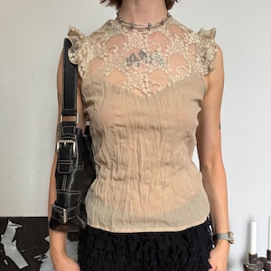 Pode incluir: Uma blusa sem mangas bege com um jugo de renda transparente com padrões florais, ombros com folhos e um corpete texturizado e enrugado. Combinada com calções de renda preta em camadas e uma mala de ombro em pele preta com fivelas.