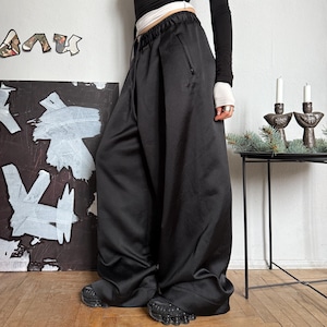 Puede incluir: Una persona con una camiseta negra de manga larga y pantalones negros de pernera ancha. Los pantalones tienen cintura con cordón y un bolsillo lateral. La persona también lleva zapatos negros. El fondo incluye una pintura y una pequeña mesa con velas.