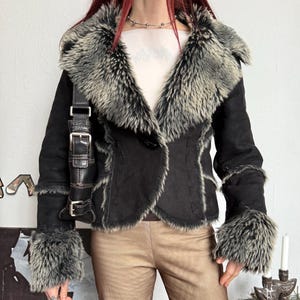 May include: Black faux fur trimmed jacket with a large collar and cuffs. The jacket has a single button closure and a suede-like texture. A black leather shoulder bag with a buckle is also visible.