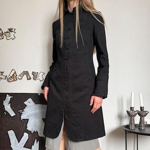 May include: A black long coat with a mandarin collar and button closures down the front. The coat has long sleeves with ruffled cuffs and a tailored fit. The coat is paired with a gray skirt and tan boots.