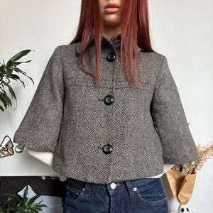 May include: A cropped, herringbone-patterned jacket in shades of gray and black, featuring three large black buttons. The jacket has a rounded collar and three-quarter sleeves. The model is wearing dark blue jeans.