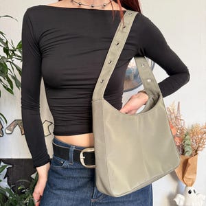May include: A sage green shoulder bag with a curved shape and a long, adjustable strap. The bag has a simple design with a smooth texture and is held by a person wearing a black long-sleeve top and blue jeans.
