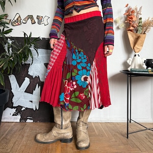 Op de afbeelding: Een veelkleurig gestreept shirt met lange mouwen en knopen, gecombineerd met een patchwork rok met bloemen, strepen en effen rode panelen. De outfit wordt gecompleteerd met beige laarzen. De rok heeft een rode tailleband.