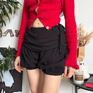 May include: A bright red cardigan with long sleeves and white buttons, worn with black gathered shorts. The shorts have a drawstring waist. The outfit is a casual, stylish ensemble.
