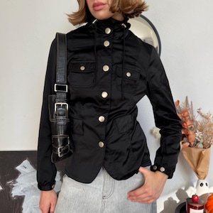 May include: A black long-sleeve jacket featuring a high collar with a drawstring, silver-tone snap buttons down the front, and two chest pockets with snap closures. The jacket has elasticized cuffs with snap details and is paired with gray jeans.