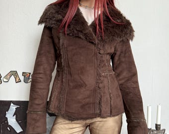 Puede incluir: Chaqueta de ante marrón con cuello y ribete de piel sintética. La chaqueta tiene una parte delantera abotonada y mangas largas. La chaqueta es de color marrón oscuro y parece ser de estilo vintage o retro.