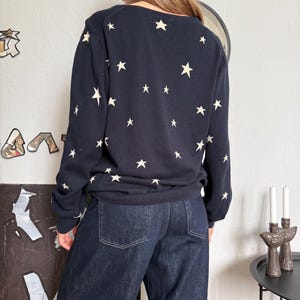以下が含まれることがあります： 背中と袖にクリーム色の星が繰り返し描かれたダークネイビーの長袖セーターと、2つのバックポケットとステッチのディテールが特徴的なダークブルーのデニムジーンズ。