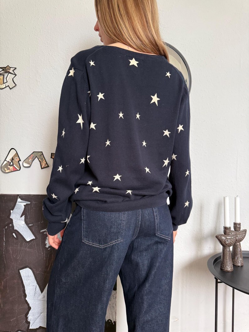 以下が含まれることがあります： 背中と袖にクリーム色の星が繰り返し描かれたダークネイビーの長袖セーターと、2つのバックポケットとステッチのディテールが特徴的なダークブルーのデニムジーンズ。