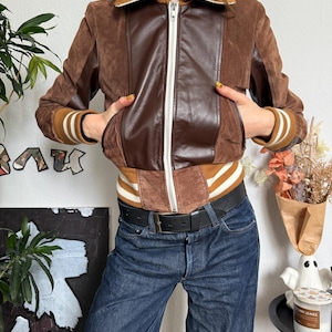 Pu&ograve; includere: Giacca bomber in pelle scamosciata e pelle marrone con cerniera bianca e polsini a righe. La giacca ha un pannello in pelle marrone sul davanti e un colletto a righe marrone e bianco. La modella indossa jeans blu e una cintura nera.