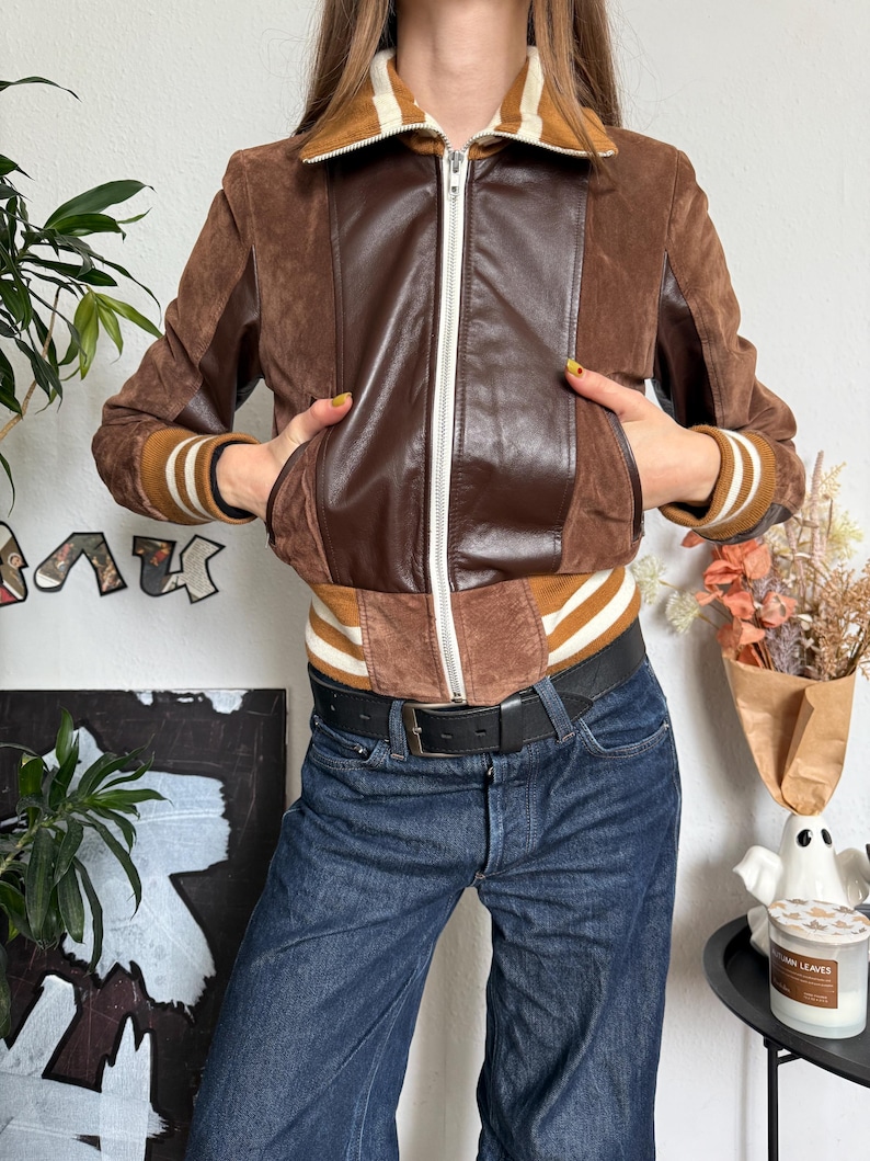 Pu&ograve; includere: Giacca bomber in pelle scamosciata e pelle marrone con cerniera bianca e polsini a righe. La giacca ha un pannello in pelle marrone sul davanti e un colletto a righe marrone e bianco. La modella indossa jeans blu e una cintura nera.