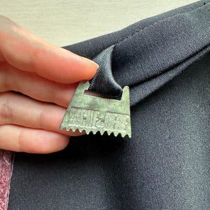 May include: Close-up of a metal picture hanger attached to a black fabric. The hanger is silver with a sawtooth edge and the text "ARCHIE & JESS" is visible. The fabric is folded over the hanger.