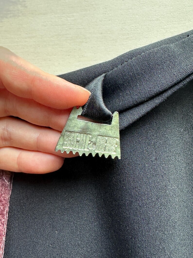 May include: Close-up of a metal picture hanger attached to a black fabric. The hanger is silver with a sawtooth edge and the text "ARCHIE & JESS" is visible. The fabric is folded over the hanger.