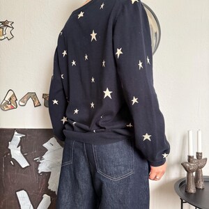 以下が含まれることがあります： 前面、背面、袖全体にクリーム色の星柄が繰り返されているネイビーブルーの長袖セーター。セーターにはリブ編みの裾と袖口があり、ポケットにステッチが見えるダークブルーのデニムジーンズと組み合わされています。