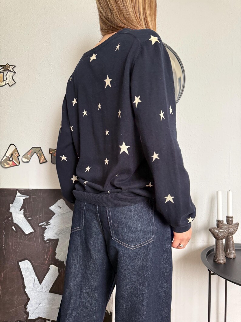 以下が含まれることがあります： 前面、背面、袖全体にクリーム色の星柄が繰り返されているネイビーブルーの長袖セーター。セーターにはリブ編みの裾と袖口があり、ポケットにステッチが見えるダークブルーのデニムジーンズと組み合わされています。