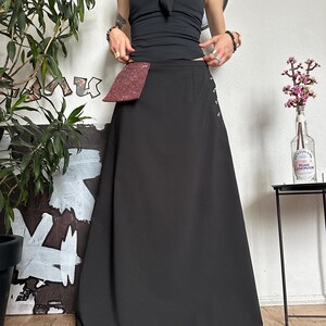 May include: A black maxi skirt with a burgundy pocket detail and side button accents. The skirt is paired with a black halter top. The outfit is styled in a minimalist aesthetic.