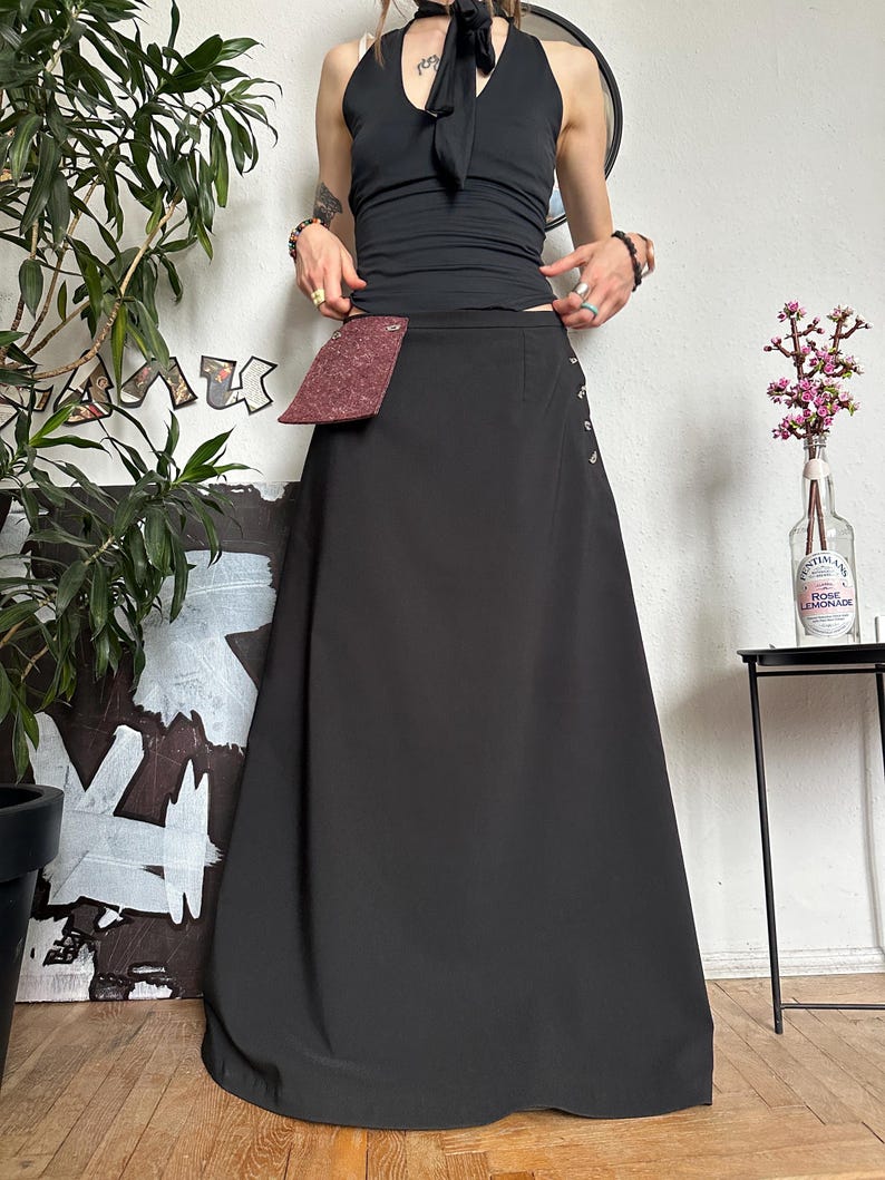 May include: A black maxi skirt with a burgundy pocket detail and side button accents. The skirt is paired with a black halter top. The outfit is styled in a minimalist aesthetic.
