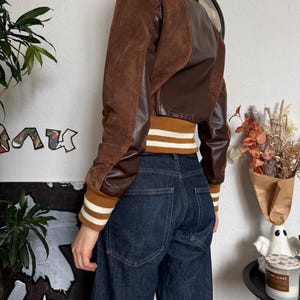 Pu&ograve; includere: Giacca bomber marrone e beige con pannelli in pelle e camoscio. La giacca ha un girovita e polsini a coste con strisce bianche. La modella indossa jeans in denim scuro.