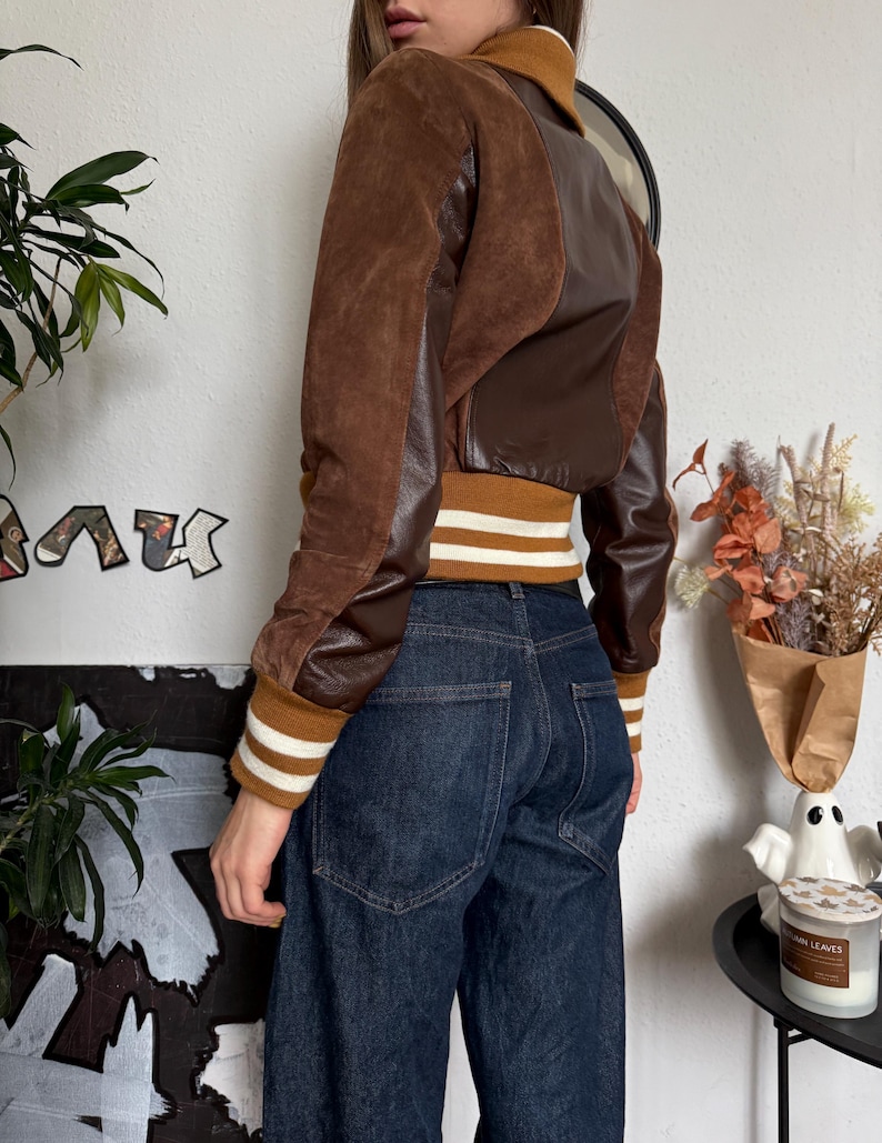 Pu&ograve; includere: Giacca bomber marrone e beige con pannelli in pelle e camoscio. La giacca ha un girovita e polsini a coste con strisce bianche. La modella indossa jeans in denim scuro.