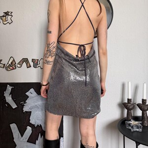 May include: A silver sequin mini dress with a halter neckline and crisscross back straps. The dress is paired with black knee-high boots. The model is seen from the back, showcasing the dress's design and fit.