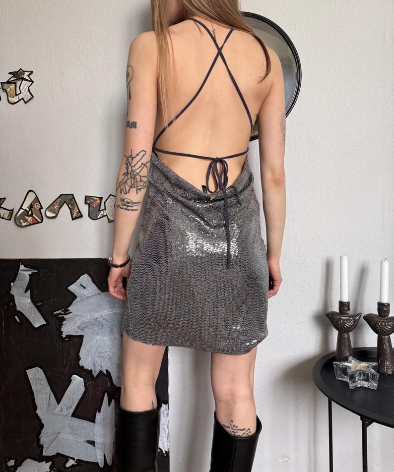 May include: A silver sequin mini dress with a halter neckline and crisscross back straps. The dress is paired with black knee-high boots. The model is seen from the back, showcasing the dress's design and fit.