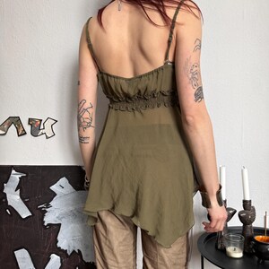 K&ouml;nnte beinhalten: Olivgr&uuml;nes Camisole-Top mit Spaghettitr&auml;gern und geraffter Empire-Taille. Das Top hat einen flie&szlig;enden, asymmetrischen Saum. Das Model tr&auml;gt eine braune Hose. Der Hintergrund ist eine wei&szlig;e Wand mit abstrakter Kunst und einem schwarzen Tisch mit Kerzen.