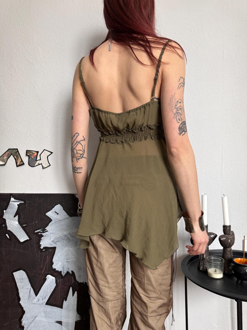 K&ouml;nnte beinhalten: Olivgr&uuml;nes Camisole-Top mit Spaghettitr&auml;gern und geraffter Empire-Taille. Das Top hat einen flie&szlig;enden, asymmetrischen Saum. Das Model tr&auml;gt eine braune Hose. Der Hintergrund ist eine wei&szlig;e Wand mit abstrakter Kunst und einem schwarzen Tisch mit Kerzen.