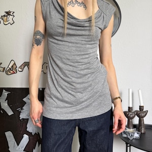 May include: A gray sleeveless top with a cowl neckline. The top has a draped design and cap sleeves. The model is wearing dark blue denim pants. The background includes wall decor and a small table with candles.