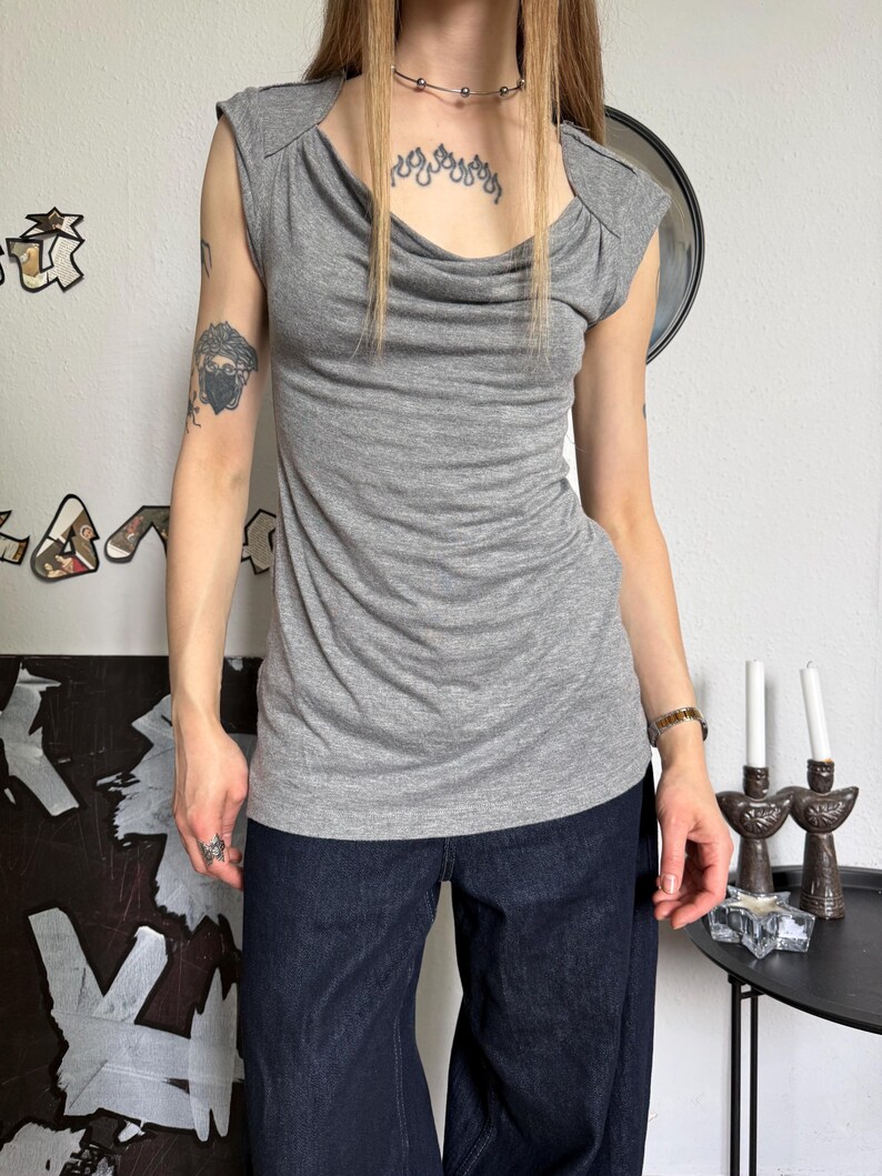May include: A gray sleeveless top with a cowl neckline. The top has a draped design and cap sleeves. The model is wearing dark blue denim pants. The background includes wall decor and a small table with candles.
