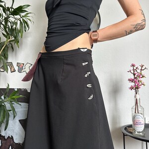 May include: A black skirt with decorative buttons and a burgundy accent. The skirt is paired with a black halter top. A watch is visible on the wrist. The image is taken indoors.