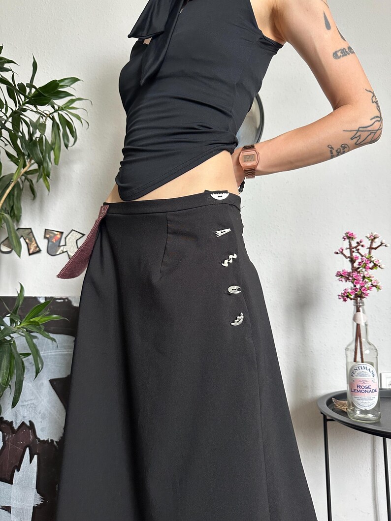 May include: A black skirt with decorative buttons and a burgundy accent. The skirt is paired with a black halter top. A watch is visible on the wrist. The image is taken indoors.