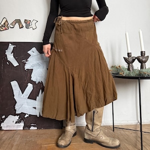 May include: A brown asymmetrical skirt with a flowing design. The skirt has a wrap-around style with a tie closure at the waist and decorative metal accents. The skirt is paired with beige boots and a black long-sleeve top.