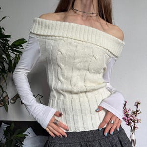 Könnte beinhalten: Cremefarbener Cable-Strickpullover mit Off-Shoulder-Ausschnitt und langen weißen Ärmeln. Der Pullover hat einen gerippten Kragen und eine taillierte Taille. Die Ärmel sind lang und eng anliegend. Das Model trägt einen grauen Rock.