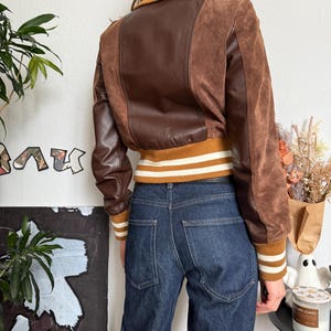 Pu&ograve; includere: Giacca bomber marrone con pannelli in pelle e camoscio. La giacca ha colletto e polsini a coste con strisce bianche. La modella indossa jeans blu scuro. La giacca &egrave; in stile vintage.