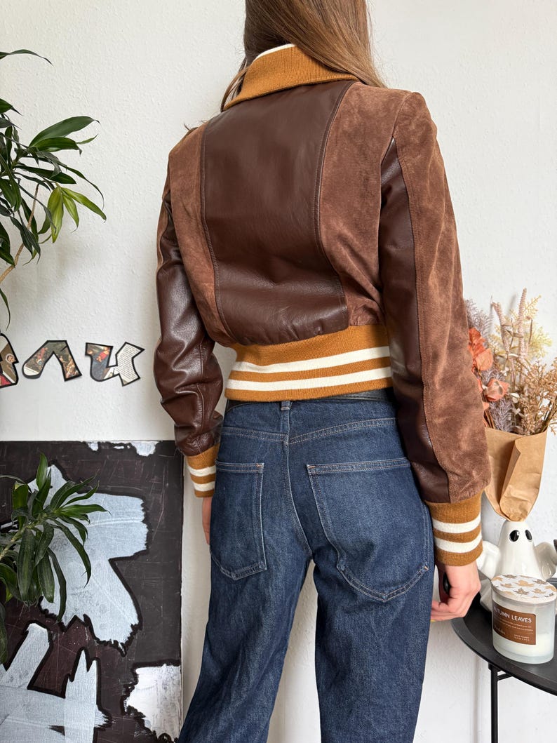 Pu&ograve; includere: Giacca bomber marrone con pannelli in pelle e camoscio. La giacca ha colletto e polsini a coste con strisce bianche. La modella indossa jeans blu scuro. La giacca &egrave; in stile vintage.