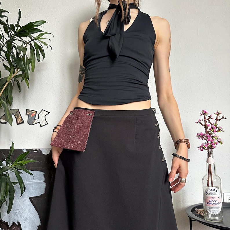 May include: A black halter top with a neck tie and a black skirt with silver buttons. The skirt has a burgundy pocket. A decorative bottle of Rose Lemonade sits on a small black table.