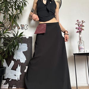 May include: A black halter top with a bow detail and a long black skirt. A small burgundy pouch is attached to the skirt. The outfit is paired with accessories and set against a white wall with a plant and artwork.