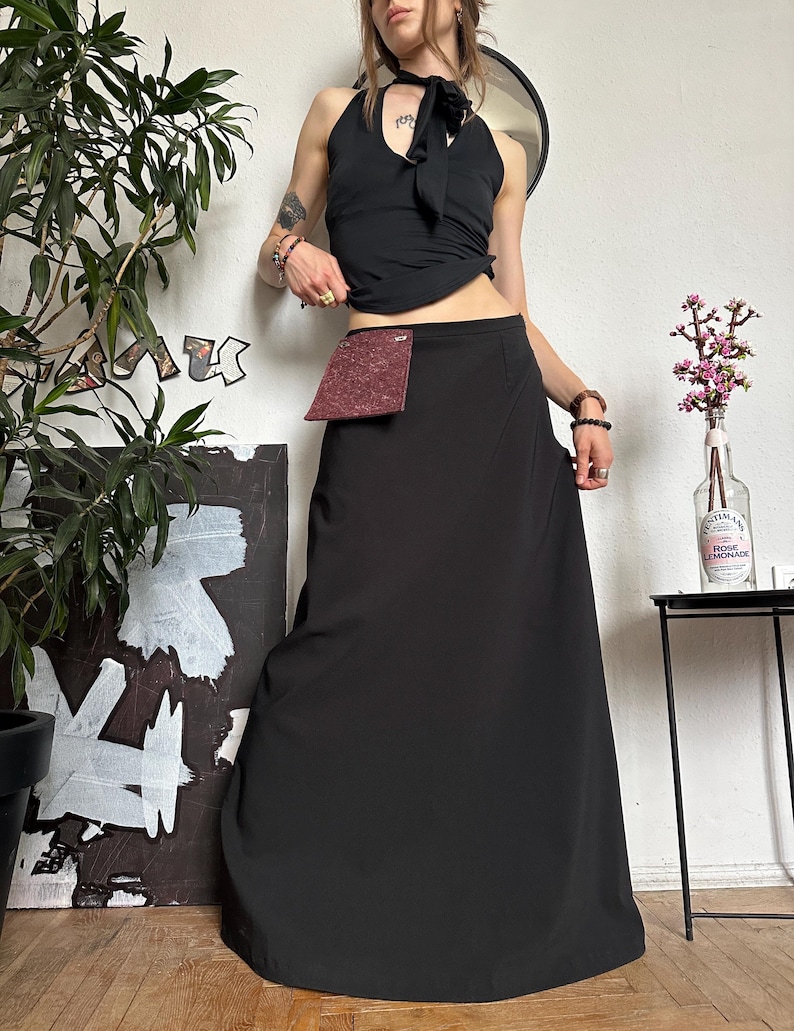 May include: A black halter top with a bow detail and a long black skirt. A small burgundy pouch is attached to the skirt. The outfit is paired with accessories and set against a white wall with a plant and artwork.