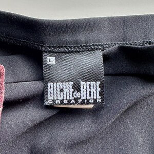 May include: Close-up of a black garment with a label. The label is rectangular, black with white text that reads "BICHE de BERE CREATION" and the size "L". The garment appears to be made of a dark, possibly synthetic fabric.