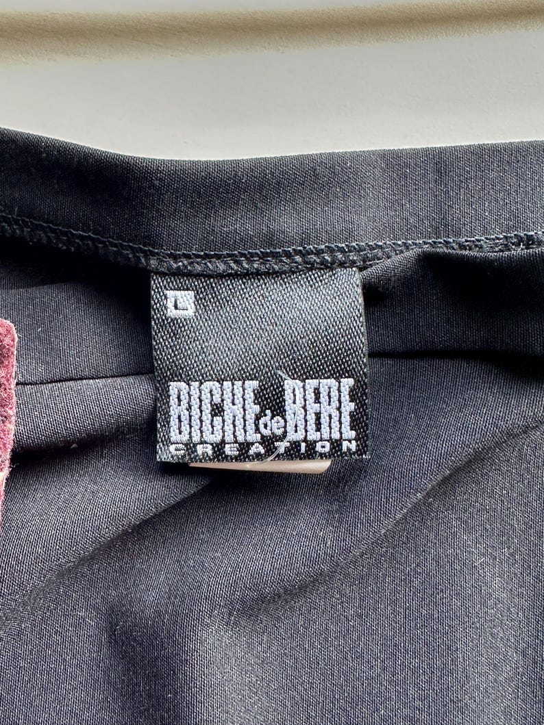 May include: Close-up of a black garment with a label. The label is rectangular, black with white text that reads "BICHE de BERE CREATION" and the size "L". The garment appears to be made of a dark, possibly synthetic fabric.