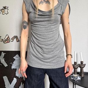 May include: A gray, sleeveless top with a draped cowl neckline. The top has a fitted silhouette and shoulder detailing. The model is wearing dark blue wide-leg jeans. The background is a plain white wall.