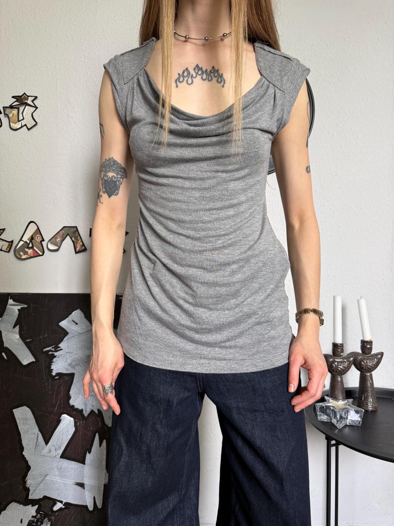 May include: A gray, sleeveless top with a draped cowl neckline. The top has a fitted silhouette and shoulder detailing. The model is wearing dark blue wide-leg jeans. The background is a plain white wall.
