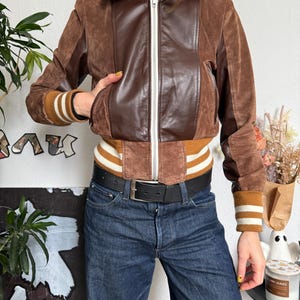 Pu&ograve; includere: Giacca bomber in pelle scamosciata e pelle marrone con cerniera bianca e polsini a righe. La giacca ha un colletto marrone e un cinturino a righe marrone e bianco. La persona indossa jeans blu scuro e una cintura nera.